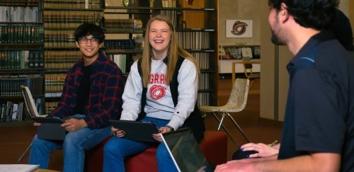 Students sitting in library with computers laughing and studying.